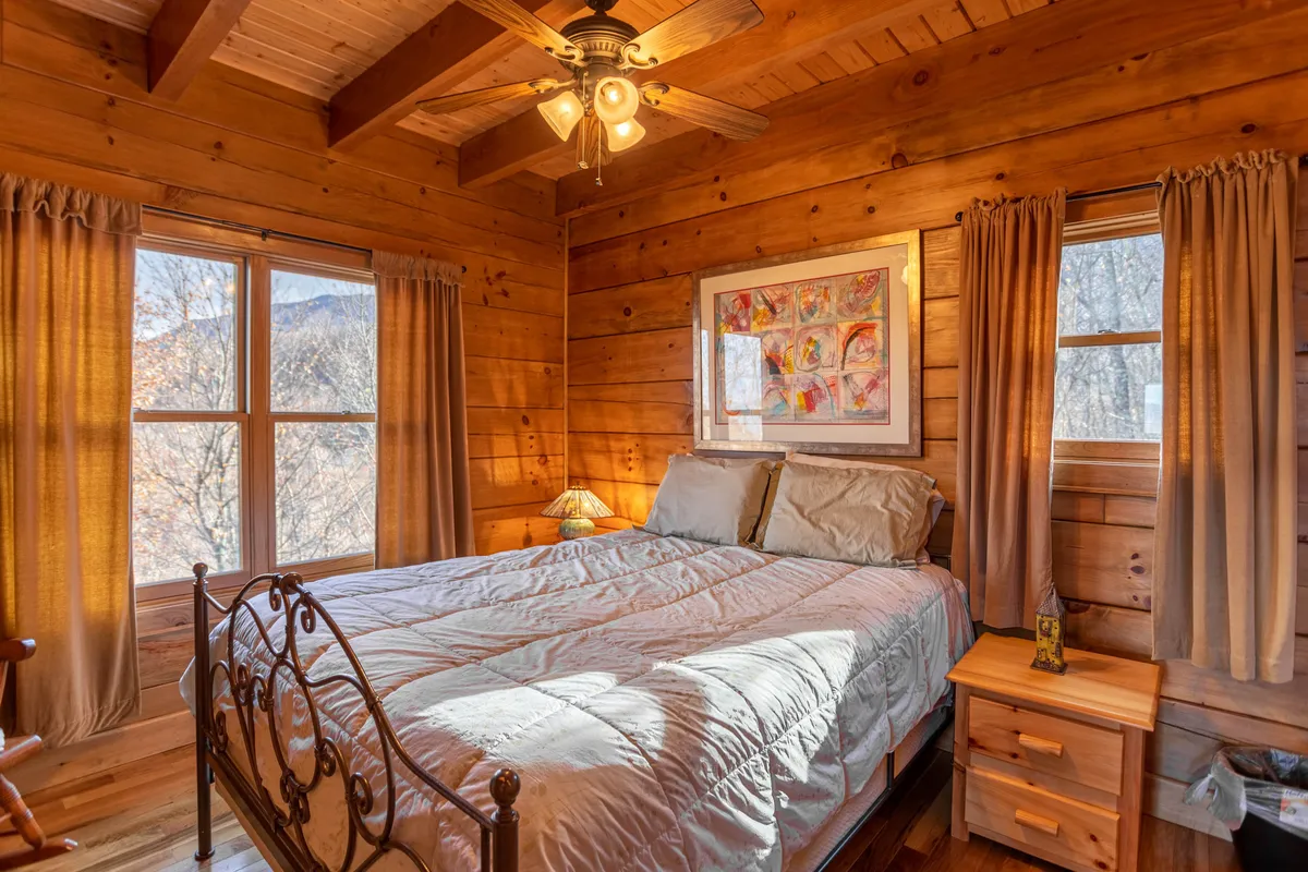 A serene bedroom with a wooden bed frame, colorful artwork on the wall, a table lamp for warm lighting, and large windows draped with curtains. The atmosphere is peaceful and inviting, perfect for unwinding after a day outdoors.