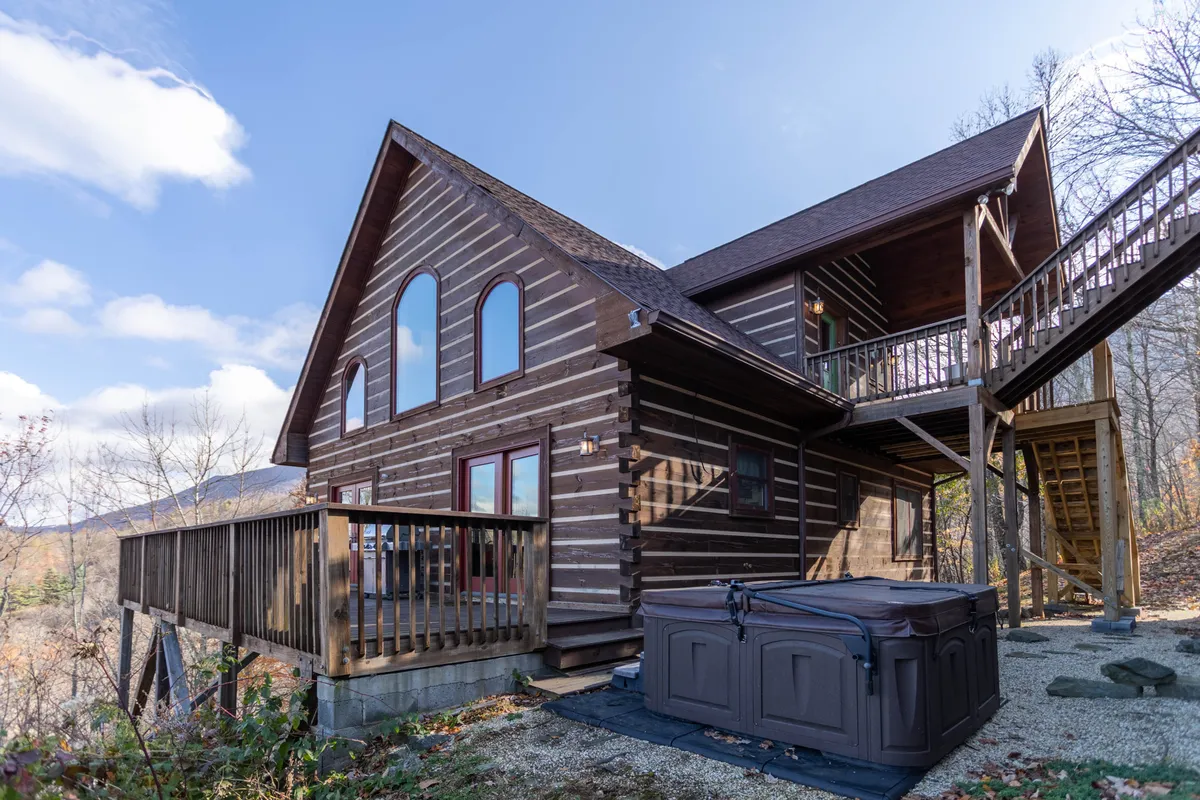 The hero image showcases a charming log cabin nestled amidst the rolling Blue Ridge Mountains, with a spacious deck featuring a private hot tub. The scene exudes tranquility, highlighting expansive 100-mile mountain views and a cozy outdoor area perfect for relaxation or stargazing.