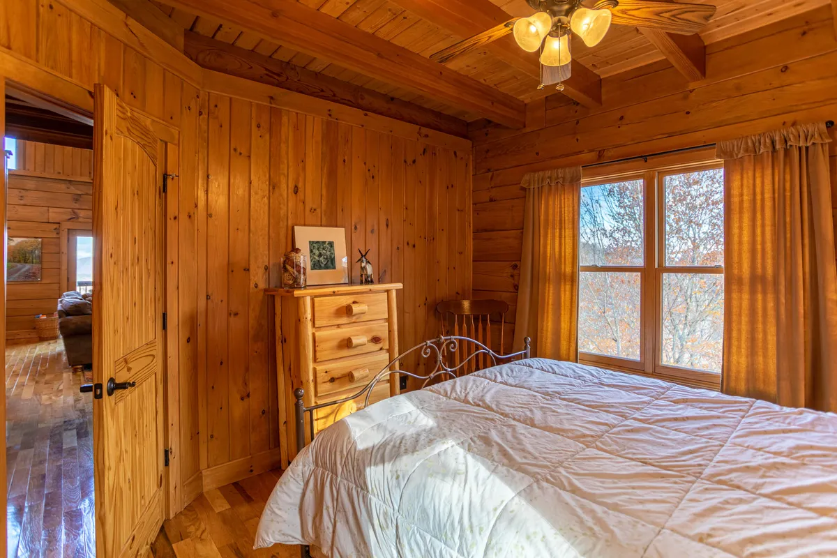 A tranquil bedroom scene with wooden walls, a cozy bed topped with a quilt, a window letting in natural light, and a chest of drawers. The space feels intimate and perfect for relaxation and a good night’s sleep.