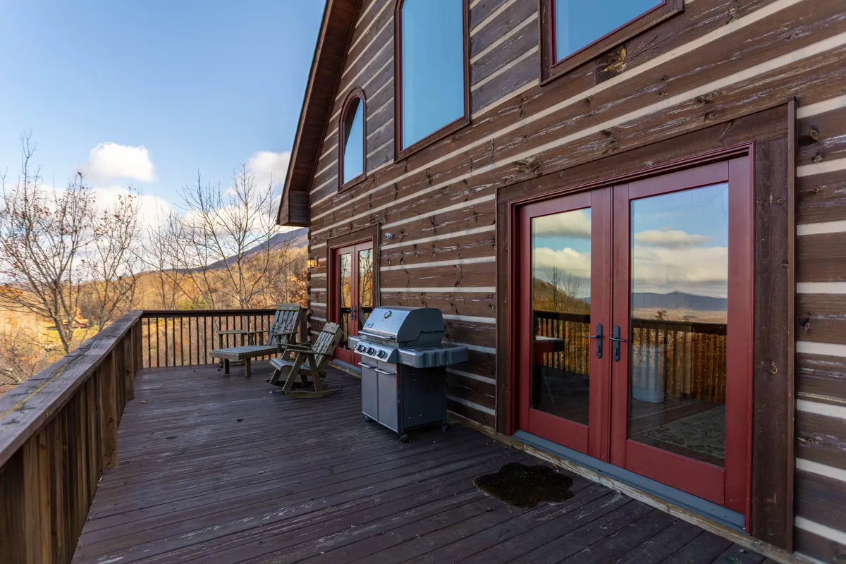 A tranquil exterior view featuring a wooden deck with comfortable outdoor seating and a grill. Large windows connect the deck to the home’s interior, while the natural surroundings and open sky invite guests to enjoy the fresh mountain air. The setup is perfect for lounging, dining outdoors, or sipping drinks while watching the sunset.