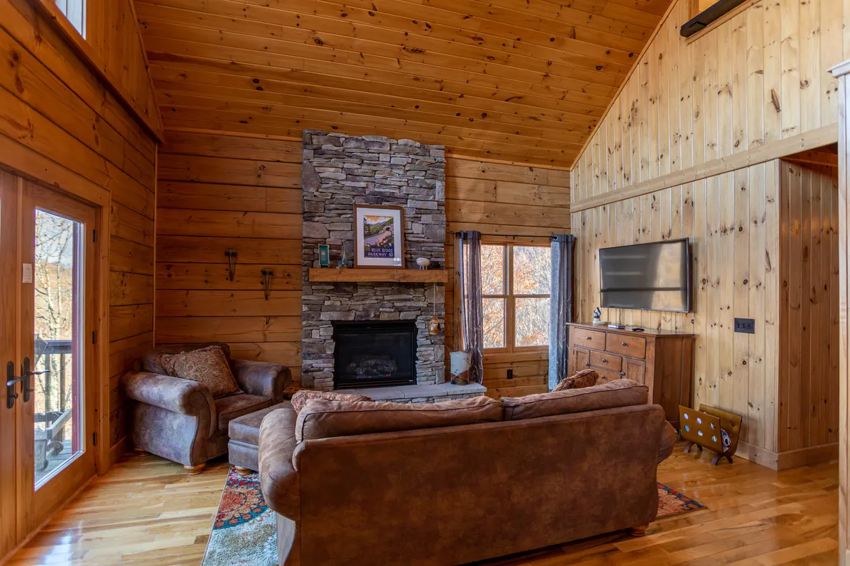 The cozy living area centers around a striking stone fireplace, surrounded by plush seating and large windows that reveal more stunning mountain vistas. The wooden walls enhance the warmth and rustic charm, making it a perfect spot for family movie nights or fireside conversations.