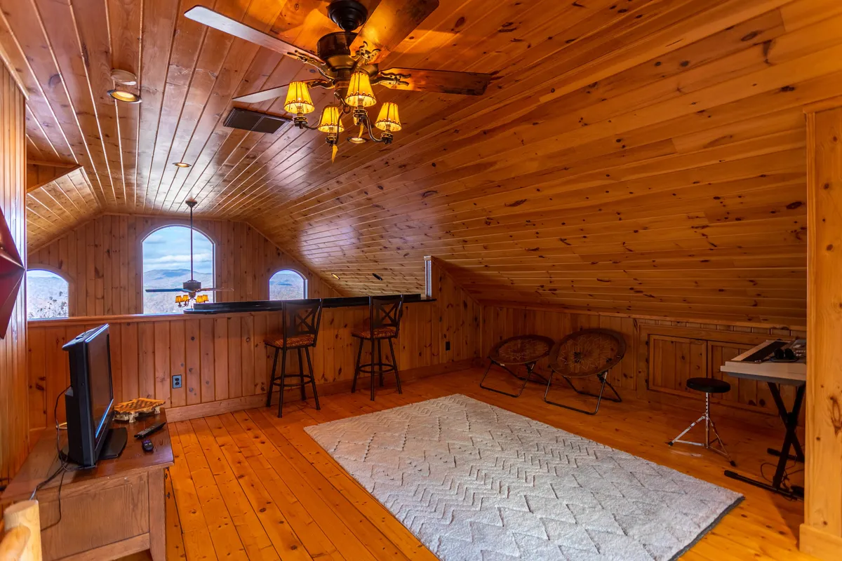 Featuring a warmly lit living area with a wooden ceiling, a bar area with stools, comfortable seating, a chandelier, and an area rug, this image suggests a welcoming space for relaxation and group interaction. The presence of windows offers connections to the outside environment.