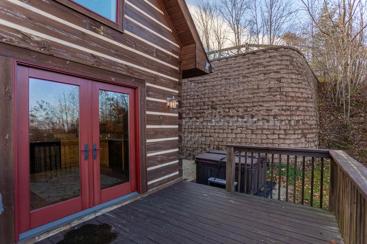 An inviting exterior scene featuring a rustic wooden deck, red double doors, and a partial view of a stone wall. The deck is an intimate spot for relaxing, socializing, or accessing the home’s hot tub. The natural setting and cozy design evoke a welcoming, pet-friendly atmosphere ideal for gatherings and quiet moments alike.
