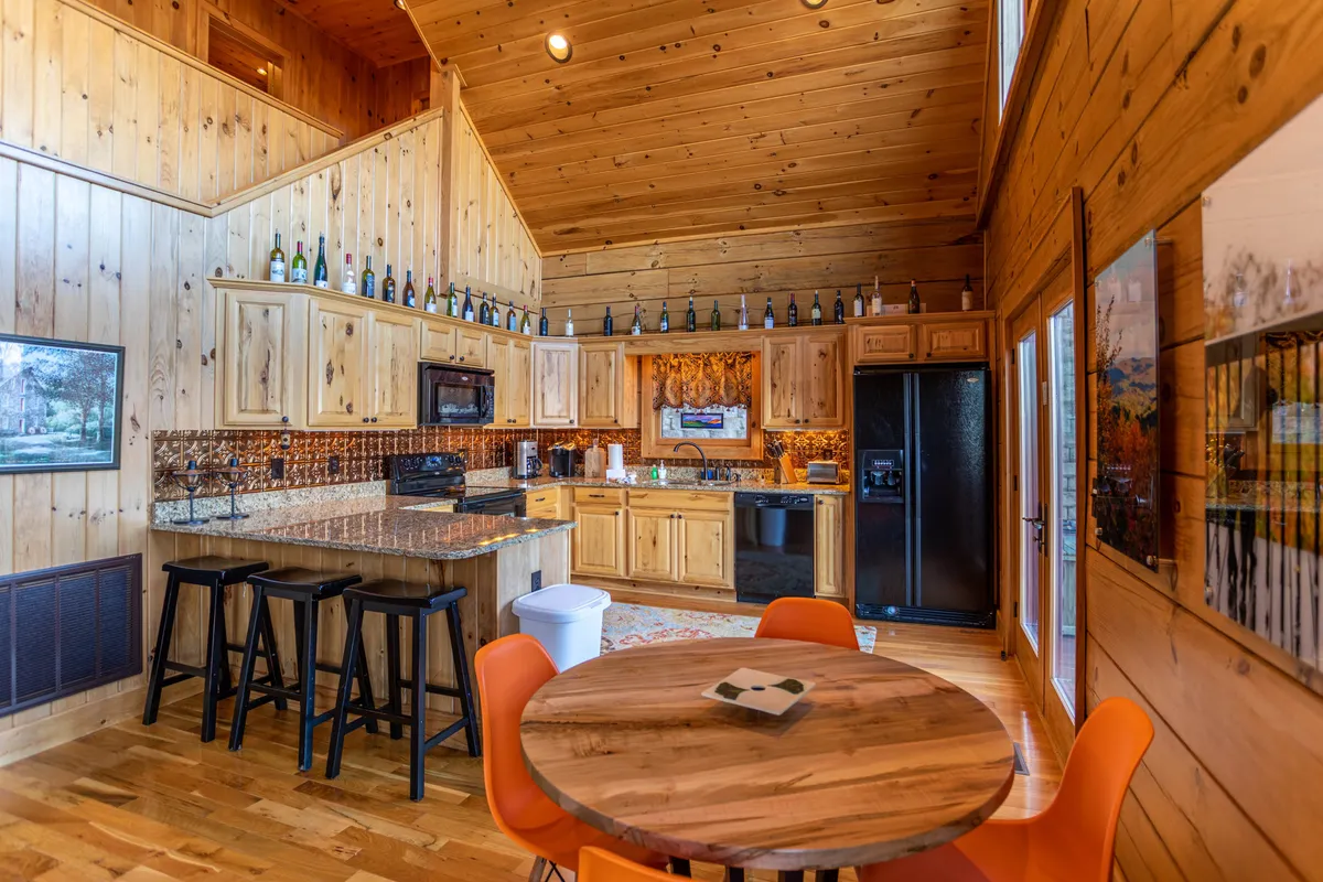 This image showcases a bright, open-plan kitchen and dining area with a kitchen island, round dining table, and large windows letting in natural light. Wooden cabinetry and bottle displays add a rustic touch, while the setup suggests a bustling hub for meals, games, and family gatherings.
