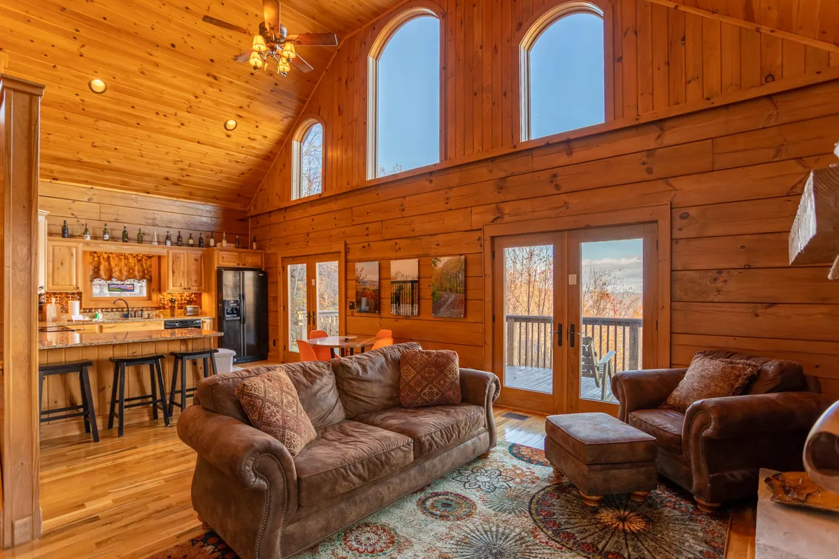A spacious open-concept living room featuring large panoramic windows that flood the area with natural light and offer sweeping views of the Blue Ridge Mountains. Warm wooden walls complement comfortable sofas and a decorative rug, while a bar area sits adjacent, perfect for entertaining. The room exudes a welcoming, communal ambiance ideal for gatherings.