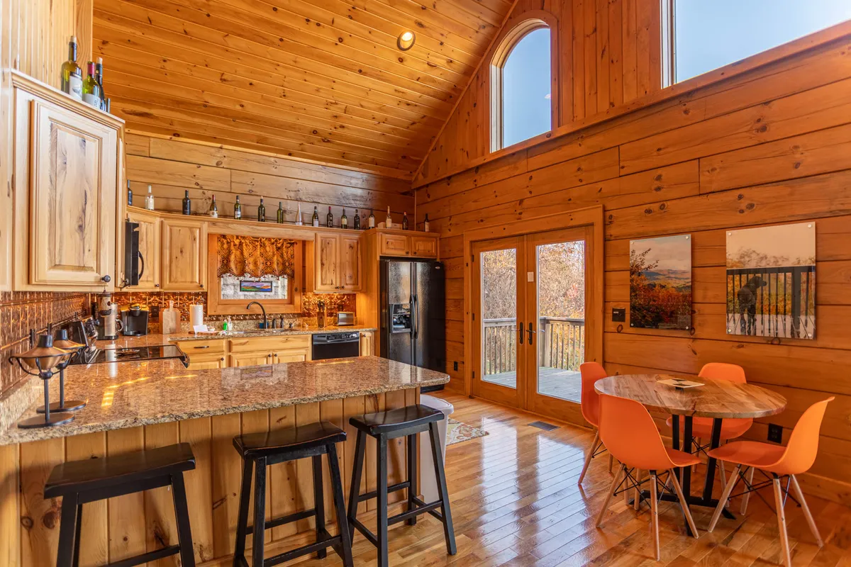 This image displays a cozy, light-filled kitchen and dining area with granite countertops, wooden cabinets, large windows, and vibrant orange dining chairs. The space is inviting, ideal for socializing, and offers a glimpse of the outdoors through expansive windows.