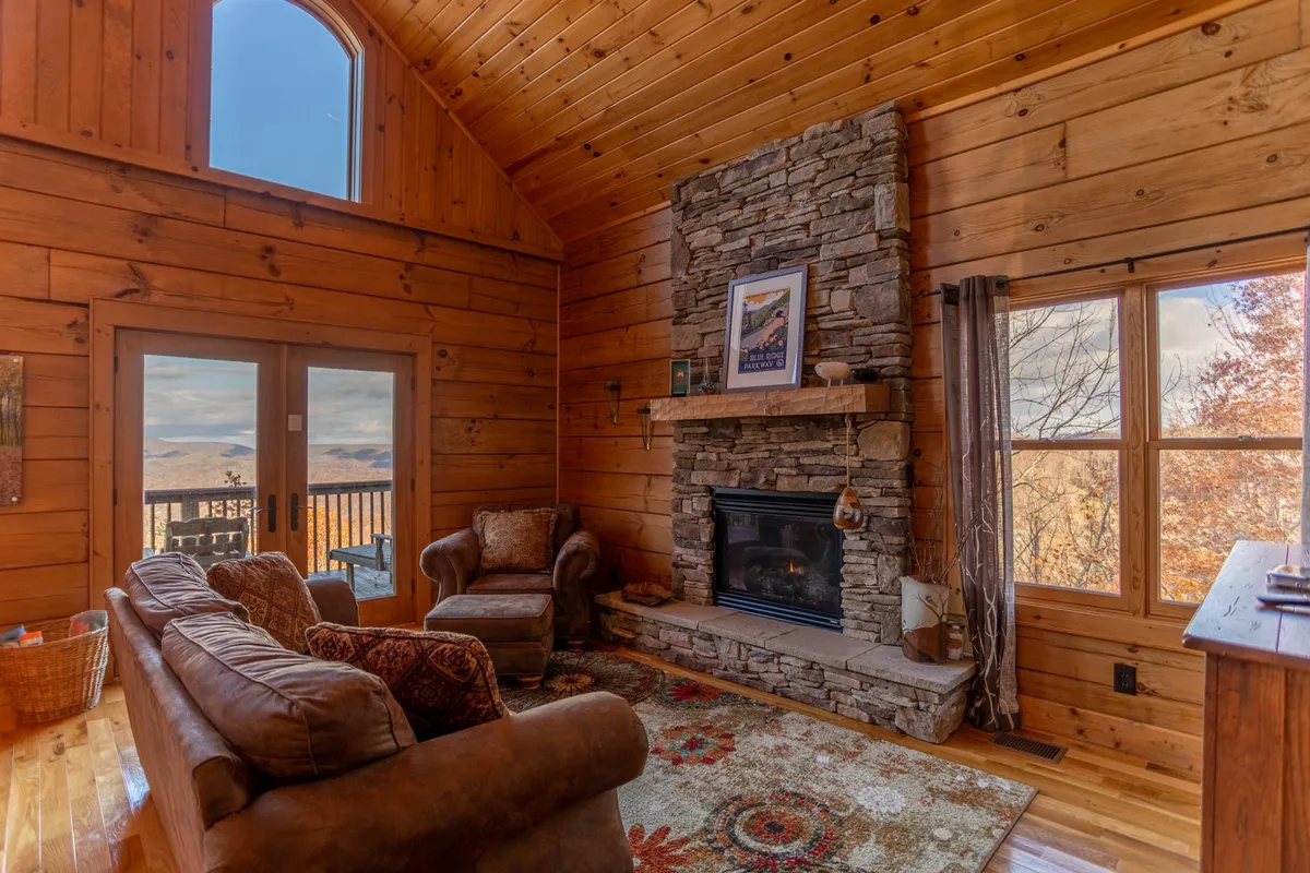 This interior image features a stone fireplace, large windows framing woodland and mountain views, rustic wooden walls, and comfortable seating. The photo conveys a sense of elevation and separation between indoor and outdoor spaces, reinforcing the mountain perch and cozy ambiance.