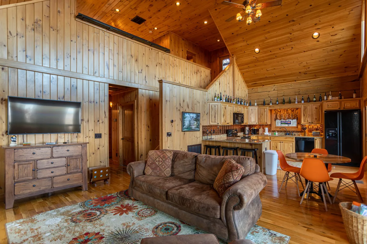 This interior image shows the main living and dining area with a comfortable brown couch, wooden dining table with orange chairs, a TV on a wooden cabinet, and a visible kitchen area. The open split-level design is implied, showcasing how the living areas are arranged for both functionality and separation.