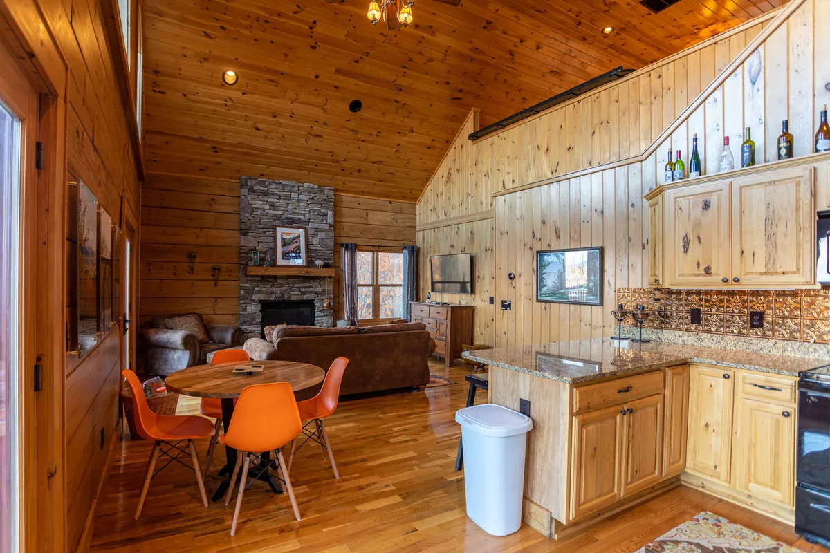 The kitchen and dining area are seamlessly integrated, featuring a wooden dining table with bold orange chairs beside a modern kitchen countertop. A stone fireplace adds warmth, inviting family and friends to cook, dine, and celebrate together in one open space.